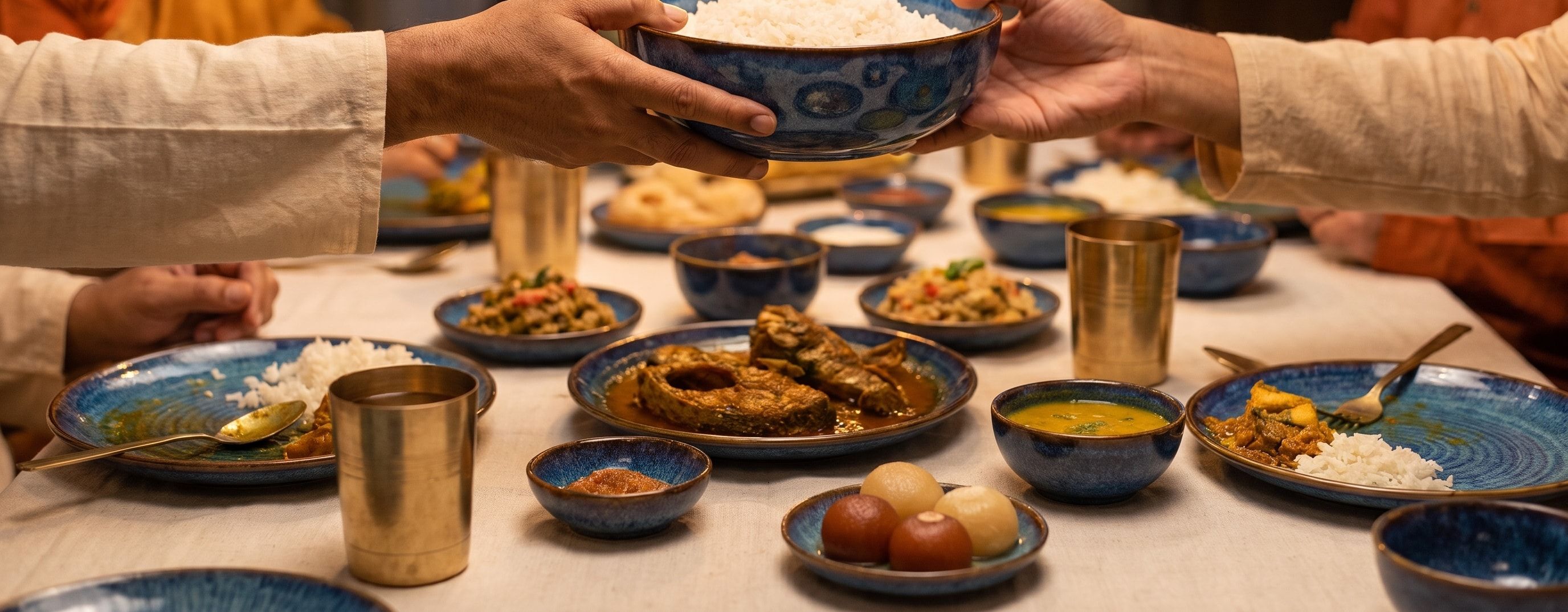 Hands passing a bowl of rice across a family table laid with blue-glazed plates, fish curry, dal and brass glasses