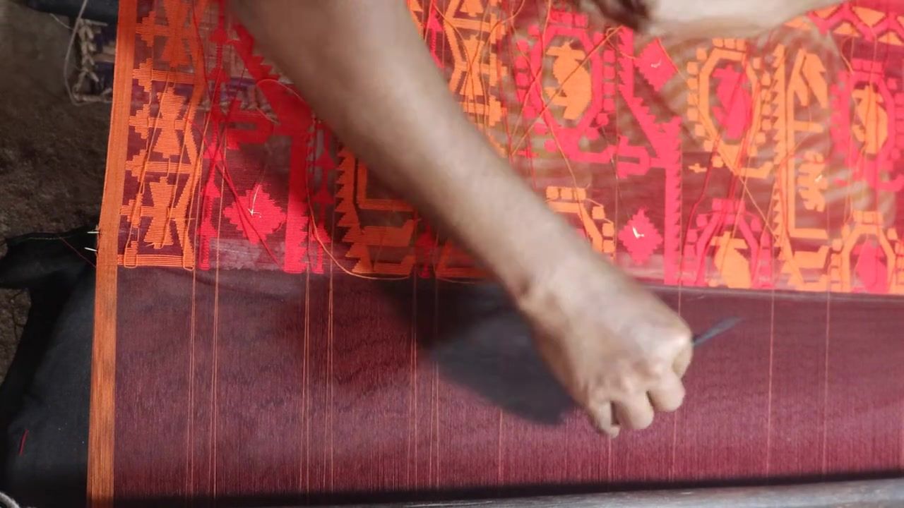 A weaver’s hand passing a shuttle across a jamdani sari on the loom, its pattern woven in orange and pink